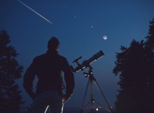 A stargazer standing next to a telescope and looking up at the sky as a meteor shoots overhead
