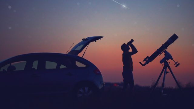 A silhouette of a man looking into the night sky with binoculars next to his car and a telescope while a meteor streaks overhead