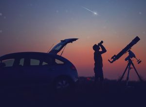 A silhouette of a man looking into the night sky with binoculars next to his car and a telescope while a meteor streaks overhead