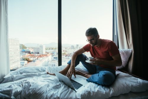 Man drinking tea and using laptop in bed