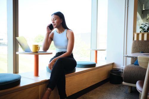 Nữ doanh nhân trẻ đang mỉm cười ngồi trên bàn trong phòng nghỉ của văn phòng và vừa nói chuyện điện thoại vừa làm việc trên máy tính xách tay