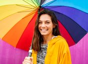 Portrait of a smiling young woman standing outside in the rain and holding a large rainbow colored umbrella