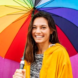 Portrait of a smiling young woman standing outside in the rain and holding a large rainbow colored umbrella