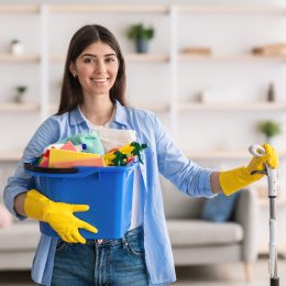 Portrait of smiling millennial lady holding bucket with cleaning supplies and mop, posing and looking at camera standing in living room.