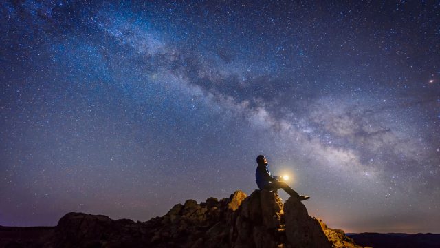 A person sitting on top of a rock outcropping with a light in their hands while looking up at the Milky Way and stars in the night sky