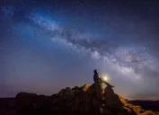 Một người ngồi trên một tảng đá bắt chéo với ngọn đèn trong tay khi nhìn lên Dải ngân hà và những vì sao trên bầu trời đêm