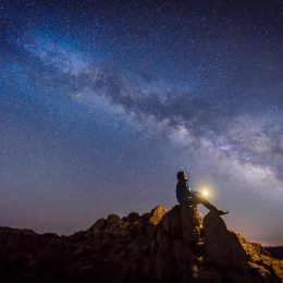 A person sitting on top of a rock outcropping with a light in their hands while looking up at the Milky Way and stars in the night sky