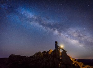 A person sitting on top of a rock outcropping with a light in their hands while looking up at the Milky Way and stars in the night sky