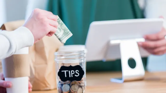 Close up of a someone putting a dollar into a tip jar next to a cash register