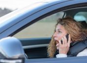 A startled looking woman using her phone to call 911 while driving