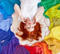 Top view of a woman with red hair laying on her bed surrounded by a circle of colorful clothes