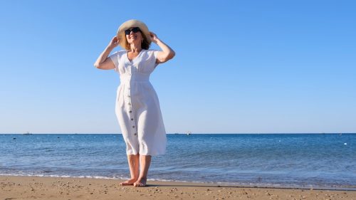 Woman stands smiling on the beach, wearing a white sundress and a straw hat