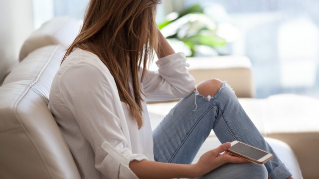 Jealous woman sitting on sofa holding phone feeling sad waiting for call, frustrated millennial girl upset or worried receiving bad news in mobile message on smartphone at home, cyberbullying concept