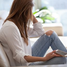 Jealous woman sitting on sofa holding phone feeling sad waiting for call, frustrated millennial girl upset or worried receiving bad news in mobile message on smartphone at home, cyberbullying concept