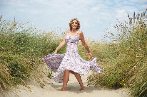 A happy middle-aged woman twirls in a patterned sundress in between sand dunes at the beach