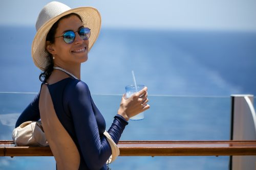 Smiling woman standing at the ocean, wearing a blue long swimsuit, beach hat and mirrored sunglasses. 
