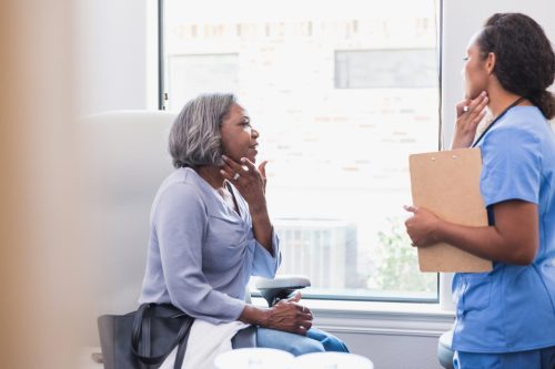 A senior woman gestures toward her face while discussing a skin rash with a female dermatologist.