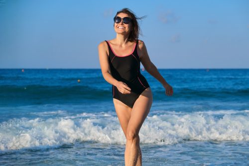 Một người phụ nữ tươi cười bước ra khỏi đại dương trong bộ đồ tắm một mảnh màu đen thể thao và đeo kính râm