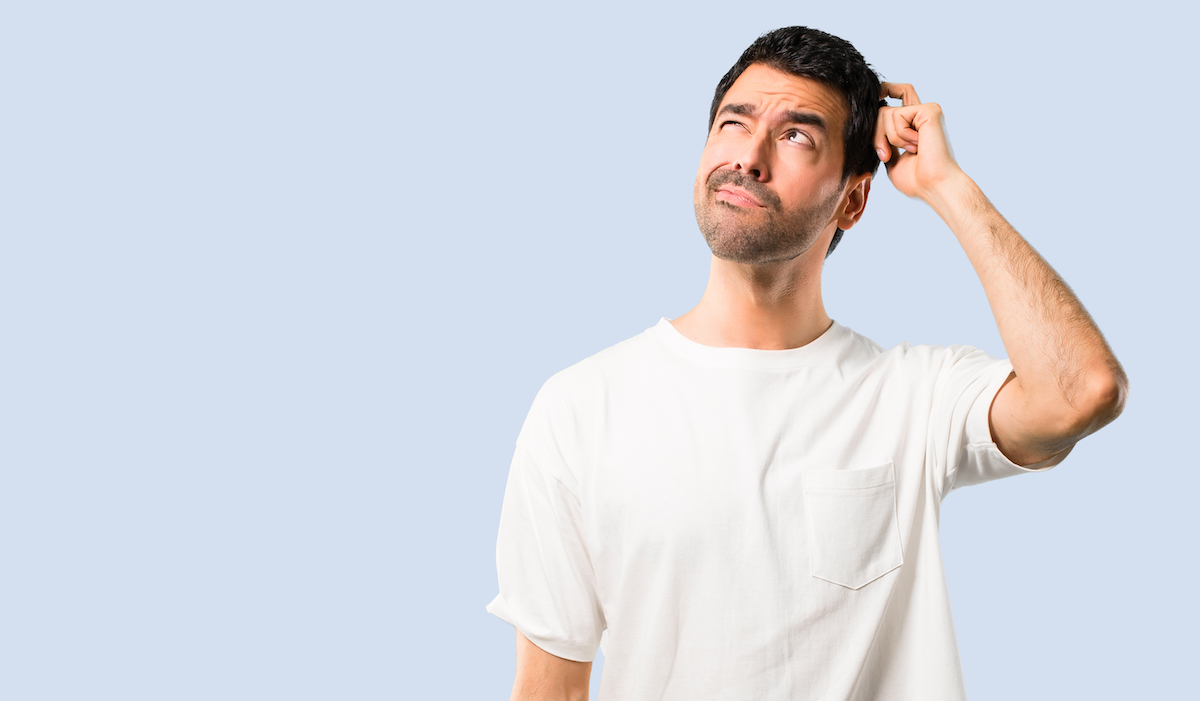 young man looking confused, scratching his head, wearing a white t-shirt against a pale blue background