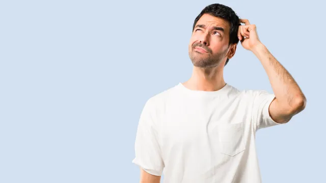 young man looking confused, scratching his head, wearing a white t-shirt against a pale blue background