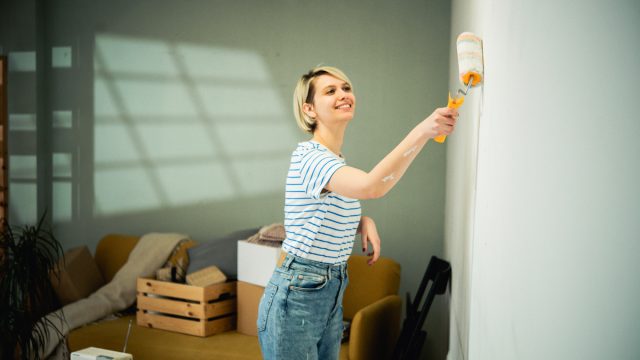 A young, smiling woman paints her walls a sage green color.