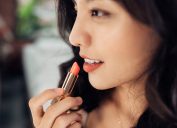 Beautiful young woman applying glossy lipstick on lips in front of a make up mirror at home