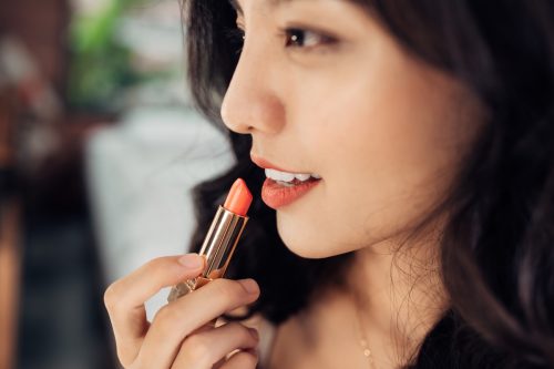 Beautiful young woman applying glossy lipstick on lips in front of a make up mirror at home