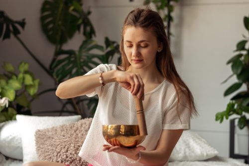 A young woman sitting at home surrounded by houseplants using a Tibetan singing bowl