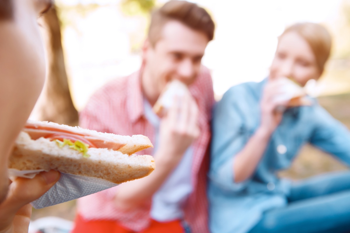 First bite. Close up of young girl eating sandwich on background of another people during picnic
