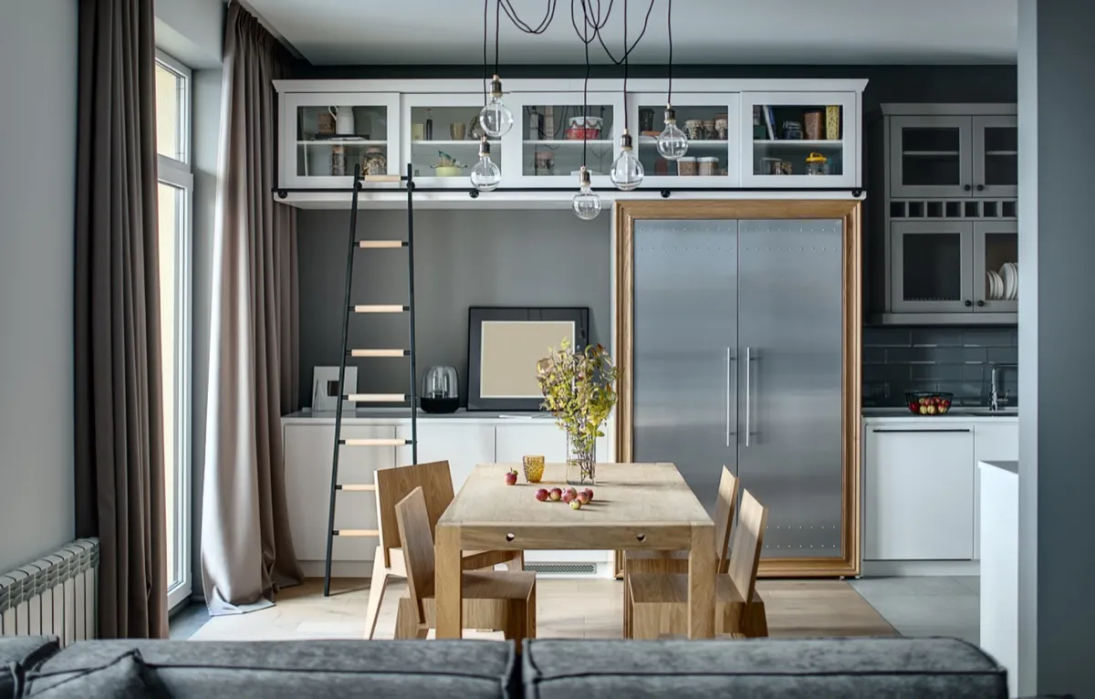Kitchen in a modern style with gray walls, white lockers and shelves with accessories. There is a wooden table with chairs, dark ladder, fridge with metal doors, sofa, sink with faucet, hanging lamps.
