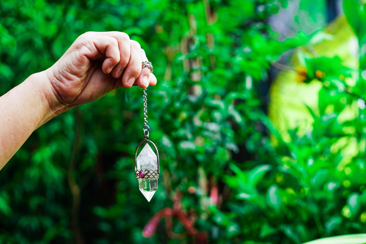 Close up of hand holding and using amethyst crystal pendulum on silver chain fortune telling pendulum in hand on nature background. The mysterious concept of the wizard.