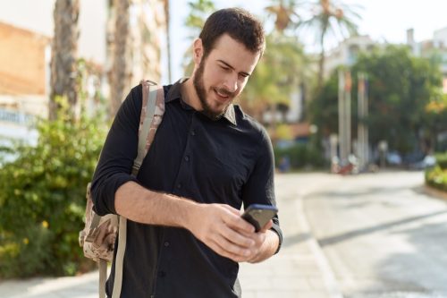 Man walking on a sidewalk with backpack, texting on his phone