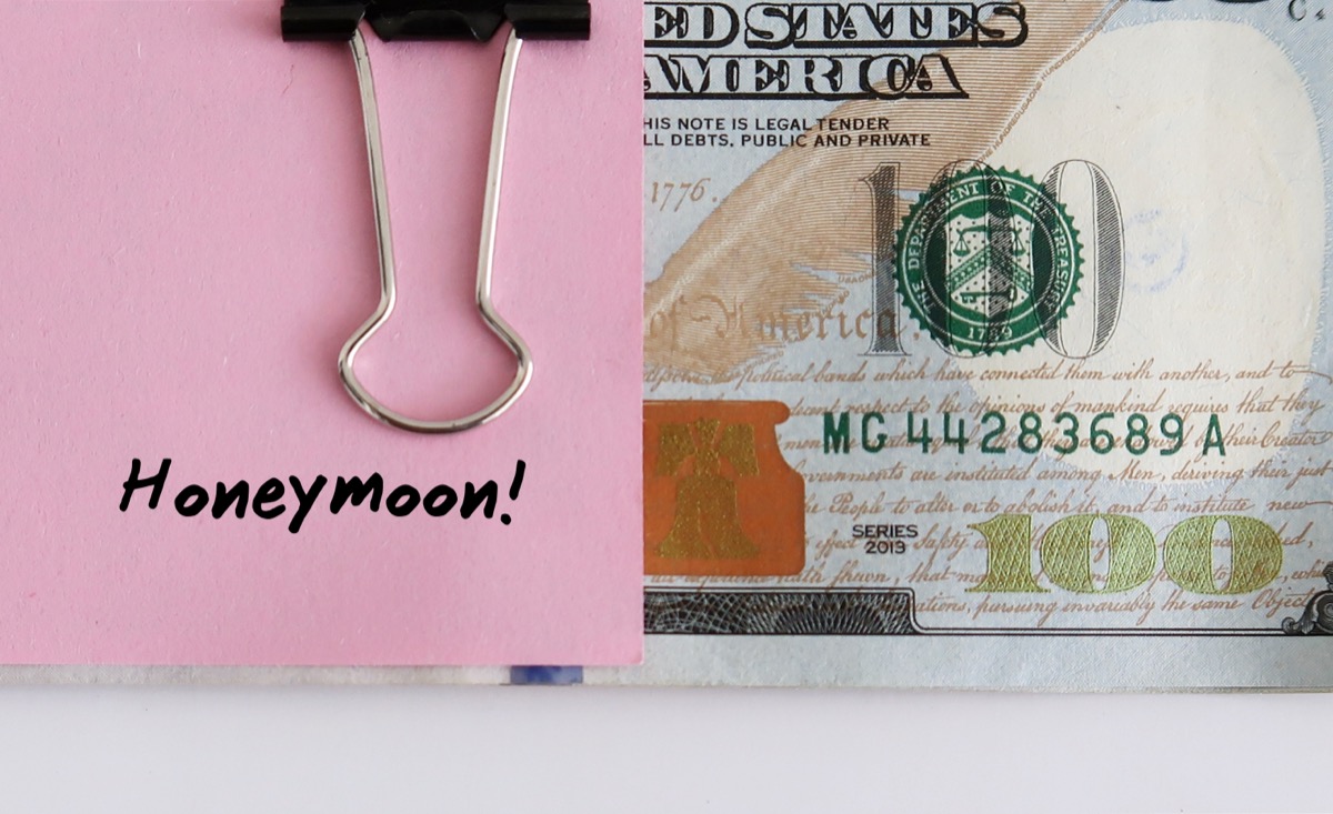 Cash money in dollars clipped with a pink note written HONEYMOON on white background - concept of saving or planning budget for honeymoon trip