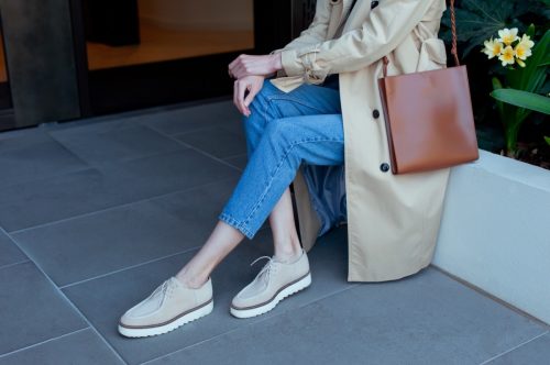 Young fashionable woman wearing beige trench coat, blue jeans and beige suede shoes. She is holding trendy brown leather handbag in hands. Street style.