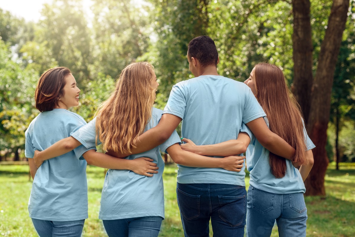 Young people girls and boy volunteers outdoors walking together in nature hugging back view talking laughing cheerful matching t-shirts