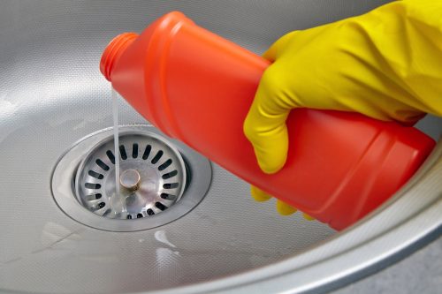 Đôi tay đeo găng tay cao su màu vàng đổ chất tẩy rửa đường ống thoát nước xuống cống thoát nước của bồn rửa bát.  Công việc làm sạch nhà bếp và cống rãnh