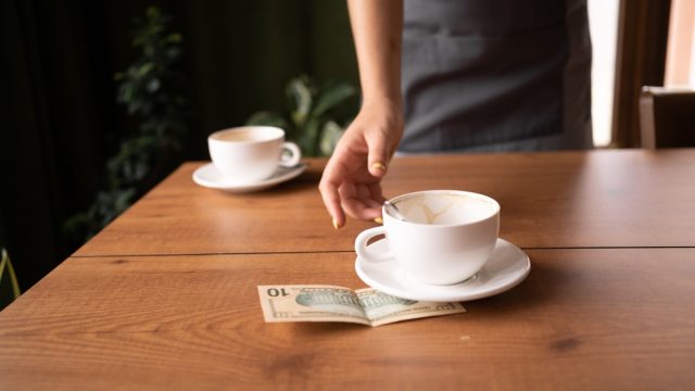 A close up of a server picking up empty coffee mugs with a $10 bill tip left under one of them.