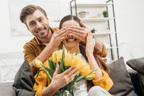happy young man covering eyes of girlfriend and presenting her bouquet of yellow tulips