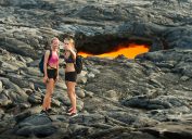 Volcanoes National Park, United States - October 17, 2016: Two young women in hiking attire stand together taking a selfie photograph in front of a glowing lava tube opening on the Big Island of Hawaii.