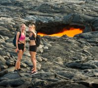 Volcanoes National Park, United States - October 17, 2016: Two young women in hiking attire stand together taking a selfie photograph in front of a glowing lava tube opening on the Big Island of Hawaii.
