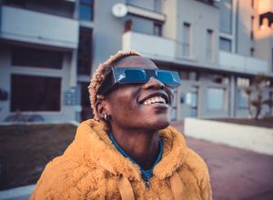 A person wearing an orange coat and protective solar glasses looking up into the sky with a smile