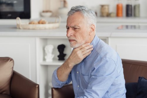 Illness and disease concept. Sick mature aged man feeling sore throat with painful swallow. Pensioner with suffering face, influenza and flu symptom sitting at home, touching neck