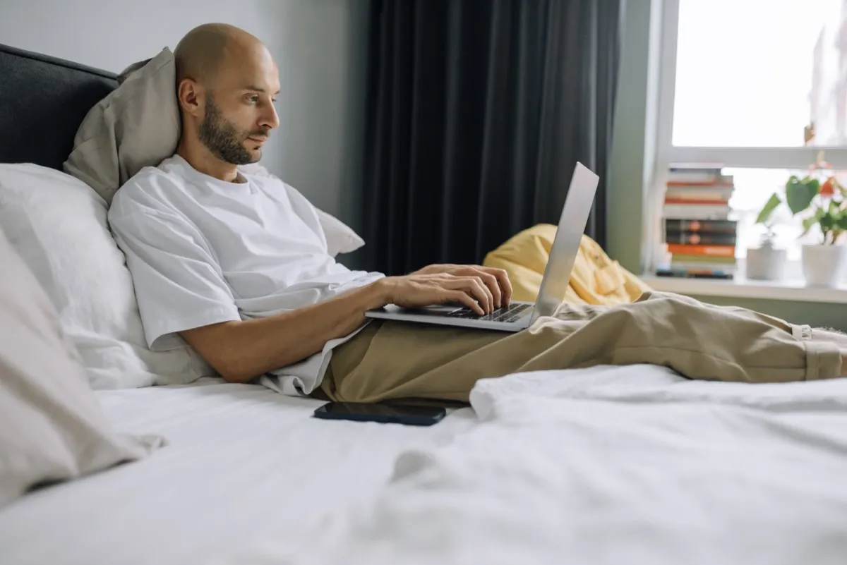 A man works on a laptop in bed in a bedroom.