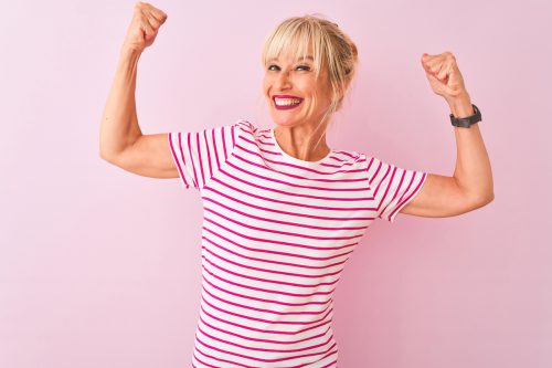 Middle age woman wearing striped t-shirt standing over isolated pink background showing arms muscles smiling proud. Fitness concept.