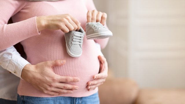 pregnant woman holding tiny shoes near her belly while husband hugs her