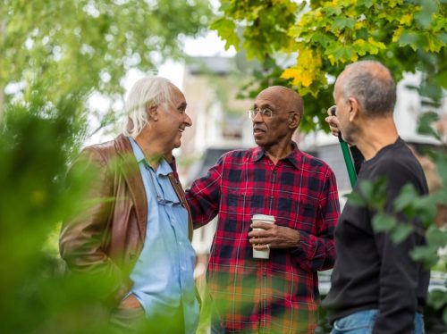 Một nhóm những người đàn ông cao cấp thuộc nhiều tầng lớp khác nhau trò chuyện trong sân trước của một người đàn ông trong khi cào lá.  Khung cảnh mùa thu rực rỡ trên đường phố một thành phố ở Bắc Mỹ.