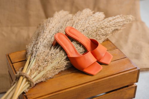 Orange mule sandals on a wooden table against a bouquet of sea grass