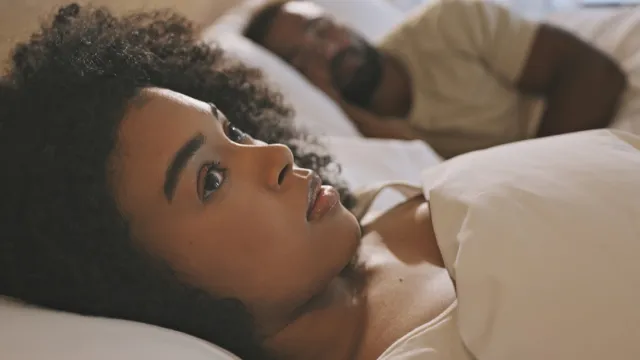 Worried female laying in bed with her husband looking anxious and concerned while thinking of her relationship issues. A man sleeping while his wife lays awake at night feeling depressed and troubled
