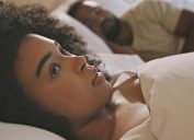 Worried female laying in bed with her husband looking anxious and concerned while thinking of her relationship issues. A man sleeping while his wife lays awake at night feeling depressed and troubled