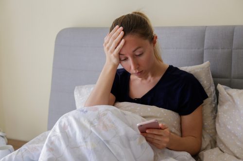 woman sitting up in bed looking at her phone with her hand on her head looking upset